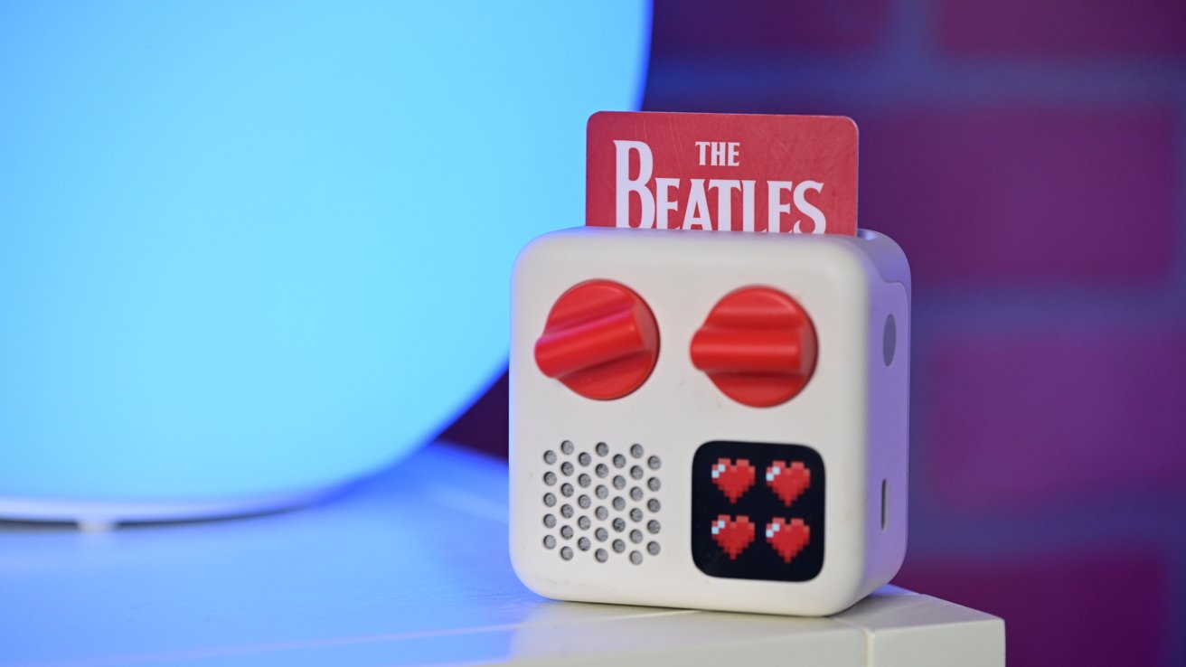 White cube device with two red dials, a pixelated heart display, and The Beatles card inserted, set against a blue and purple background.