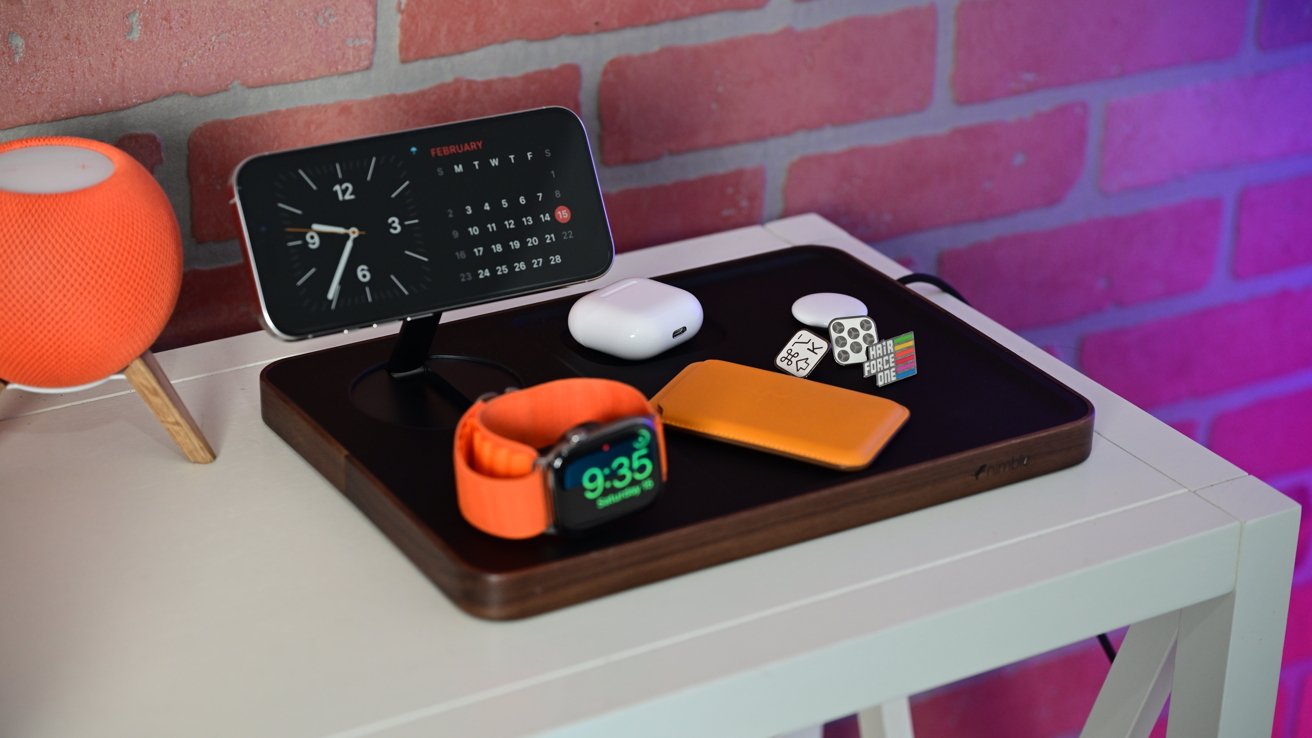 Smartwatch, clock, calendar, AirPods, orange speaker, and small accessories arranged on a white table with a brick wall background.