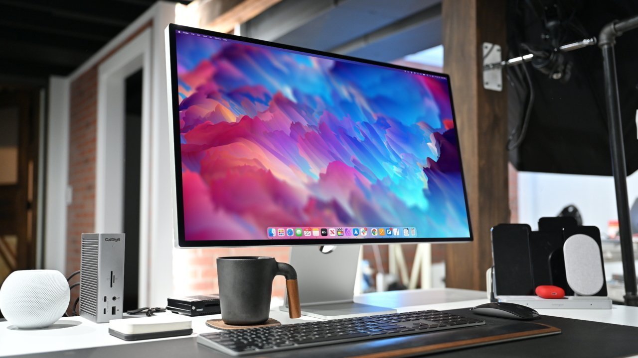 Desktop setup with large monitor displaying colorful abstract art, keyboard, mouse, black mug on coaster, smart speaker, and tech gadgets on a white desk.