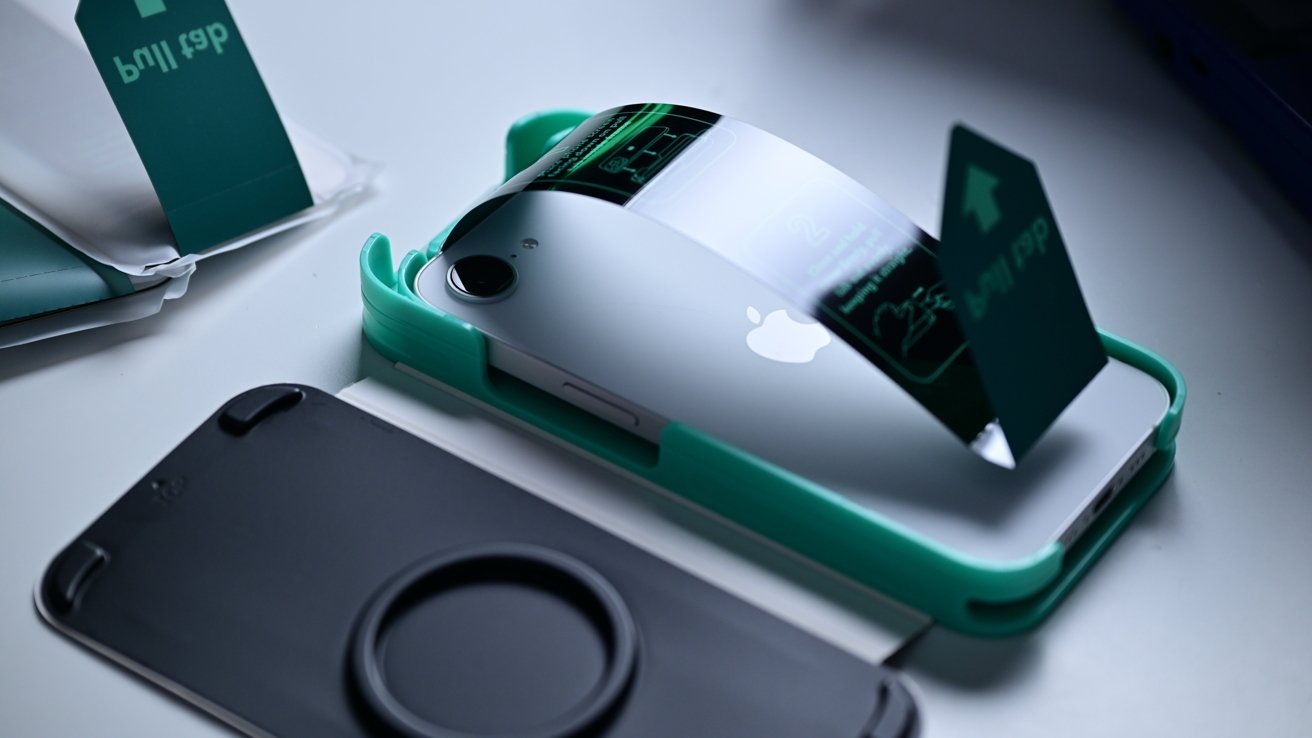 White phone with Apple logo in green stand, displaying green holographic screen, on white table next to dark circular base.