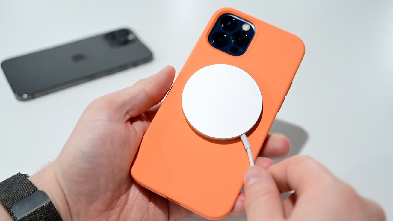 A hand holding a smartphone with an orange case attached to a circular white charger, with another phone visible in the background.