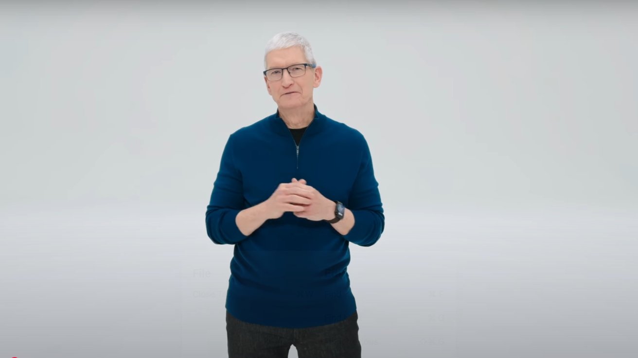 Tim Cook with short gray hair and glasses, wearing a dark blue sweater, black pants, and a watch, standing in a light-colored room.