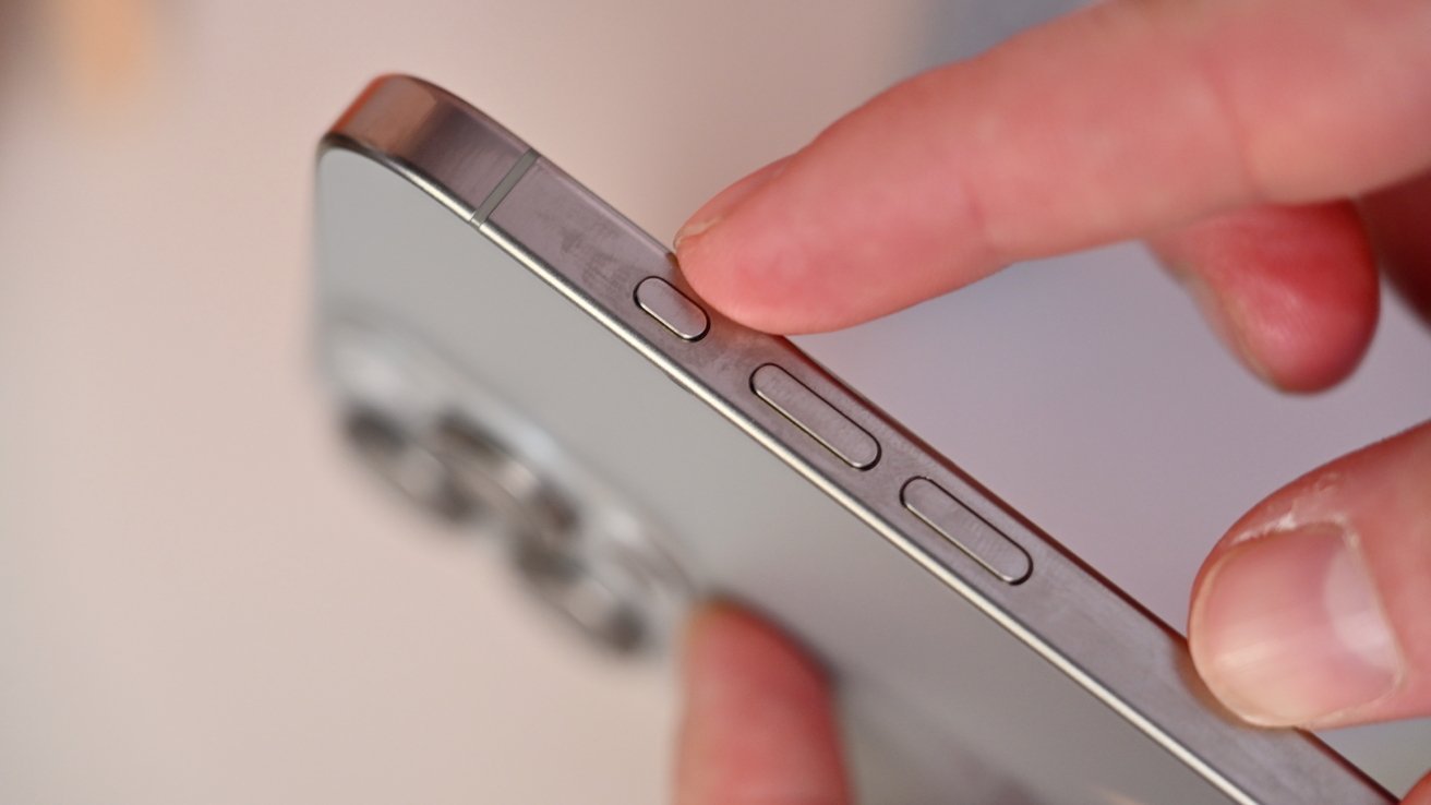 A hand presses the button on the side of a silver smartphone, with camera lenses visible in the background.