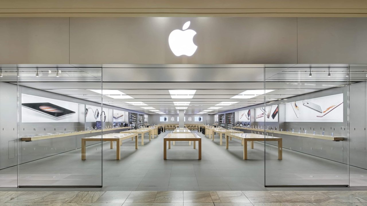 Bright, modern electronic store with glass entrance, wooden tables, illuminated displays of tablets, watches, and smartphones, featuring a glowing apple logo above.