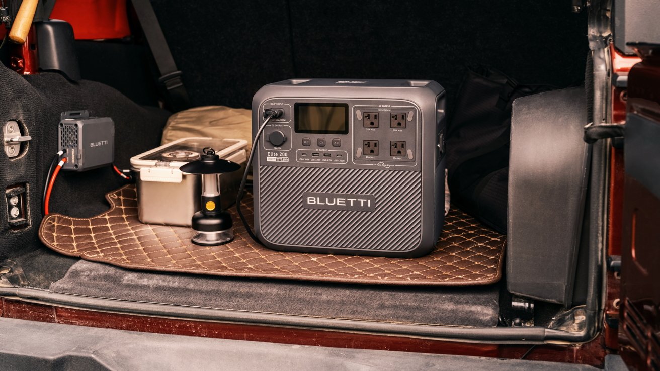 Portable power station with display and outlets sits on a cushioned mat in a vehicle trunk. Nearby are a lantern and a small box.