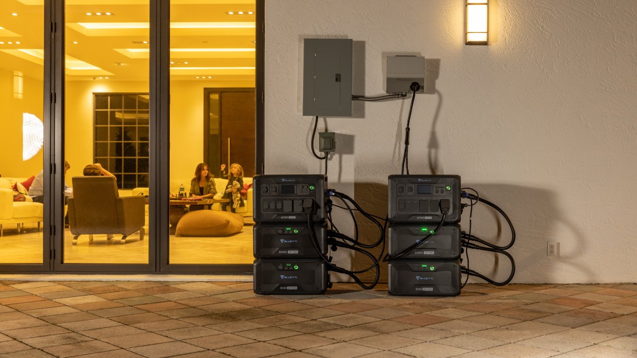 Power stations connected to a wall outside; indoors, people sitting in a warmly lit room visible through glass doors.