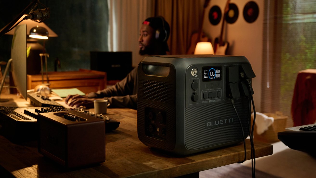 Person with headphones at a desk with computer and coffee cup, next to a large portable power station labeled Bluetti, guitars and records in the background.