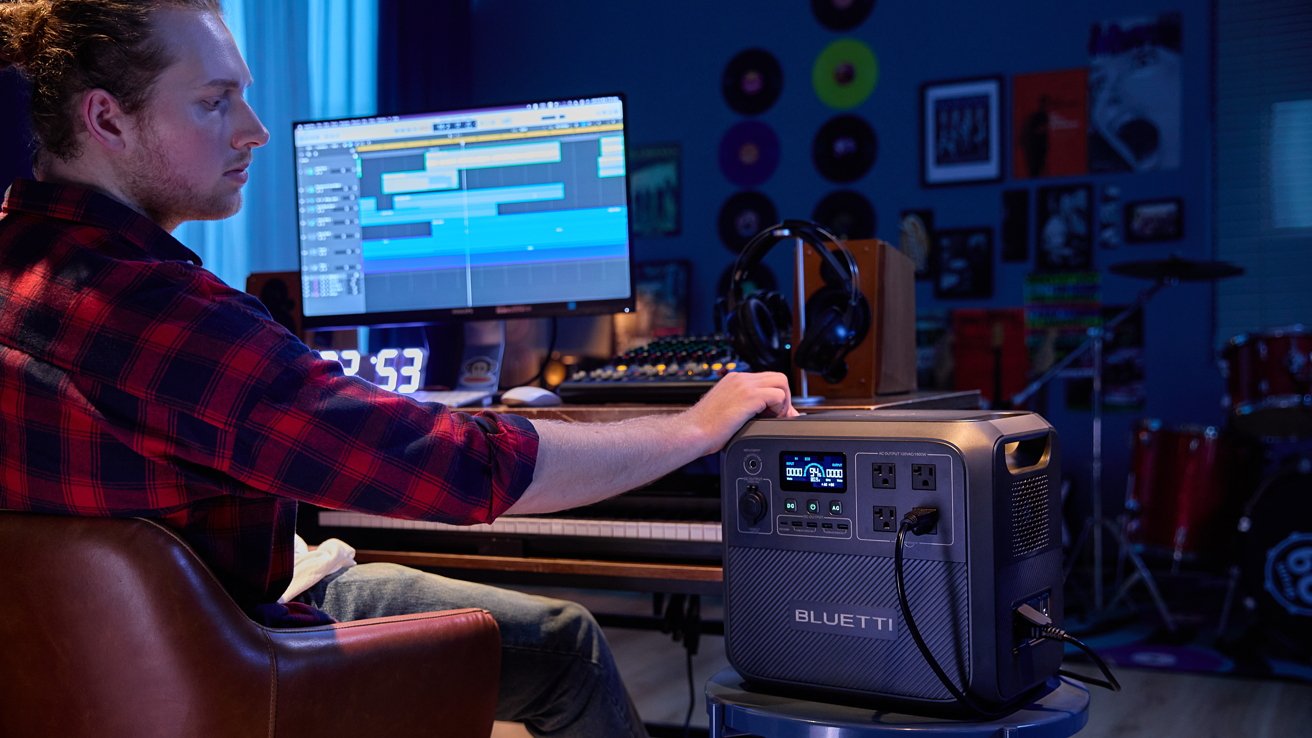 Person in a studio setting using a Bluetti power station next to a computer with music software, surrounded by recording equipment and decor.