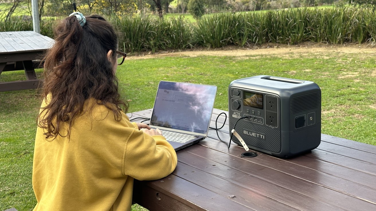 Person wearing a yellow sweater using a laptop at an outdoor wooden table with a Bluetti power station. Green grass and shrubs visible in the background.