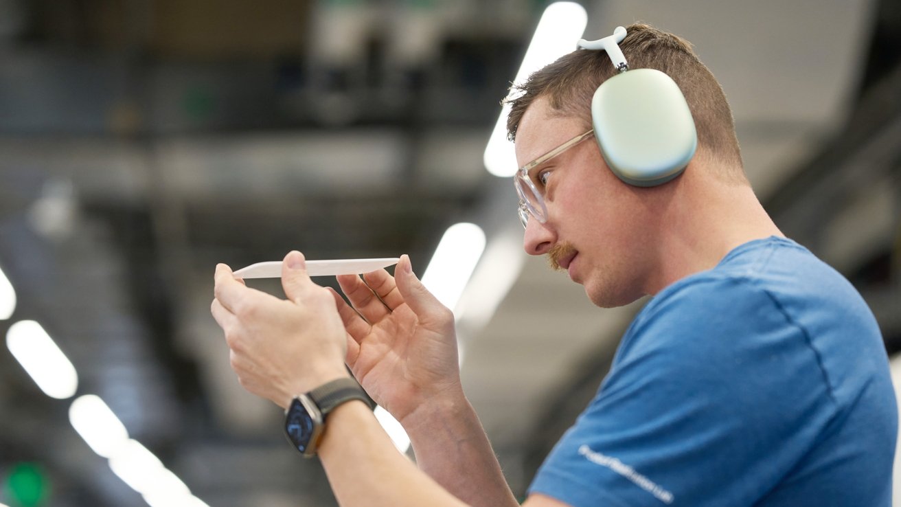 Person in blue shirt wearing headphones, closely examining a thin rectangular object, with blurred industrial background.