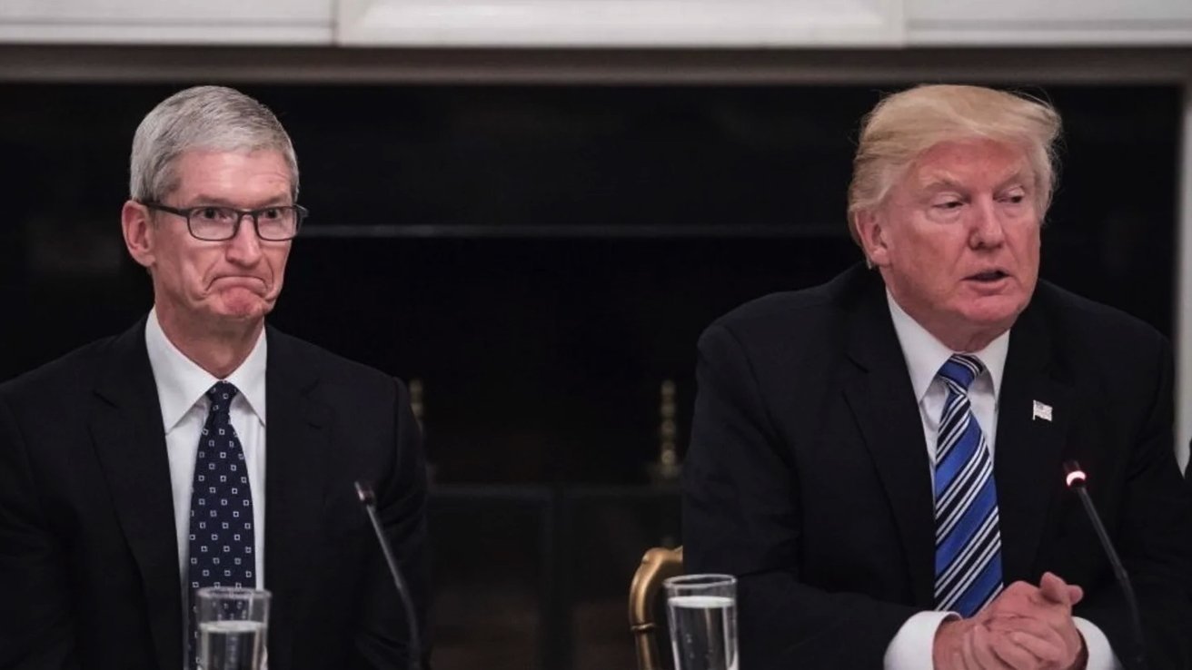 Two men in suits sit at a table. One appears thoughtful, while the other listens intently. A microphone and glasses of water are in front of them.