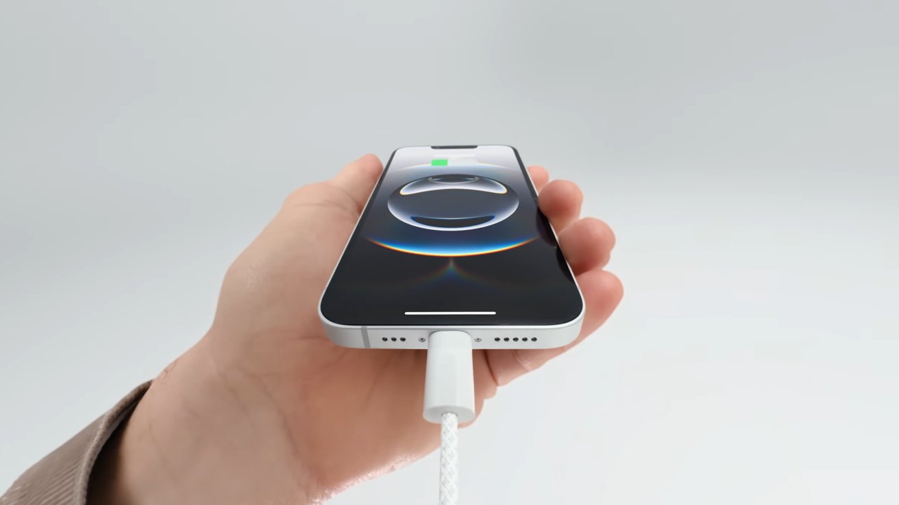 A hand holds an iPhone 16e with a charging cable connected, displaying a charging symbol on the screen against a light background.