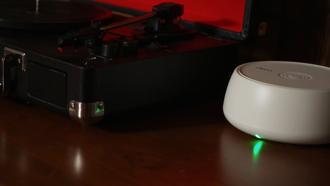 A black turntable with a red lid open next to a small white device emitting a green light, on a wooden surface.