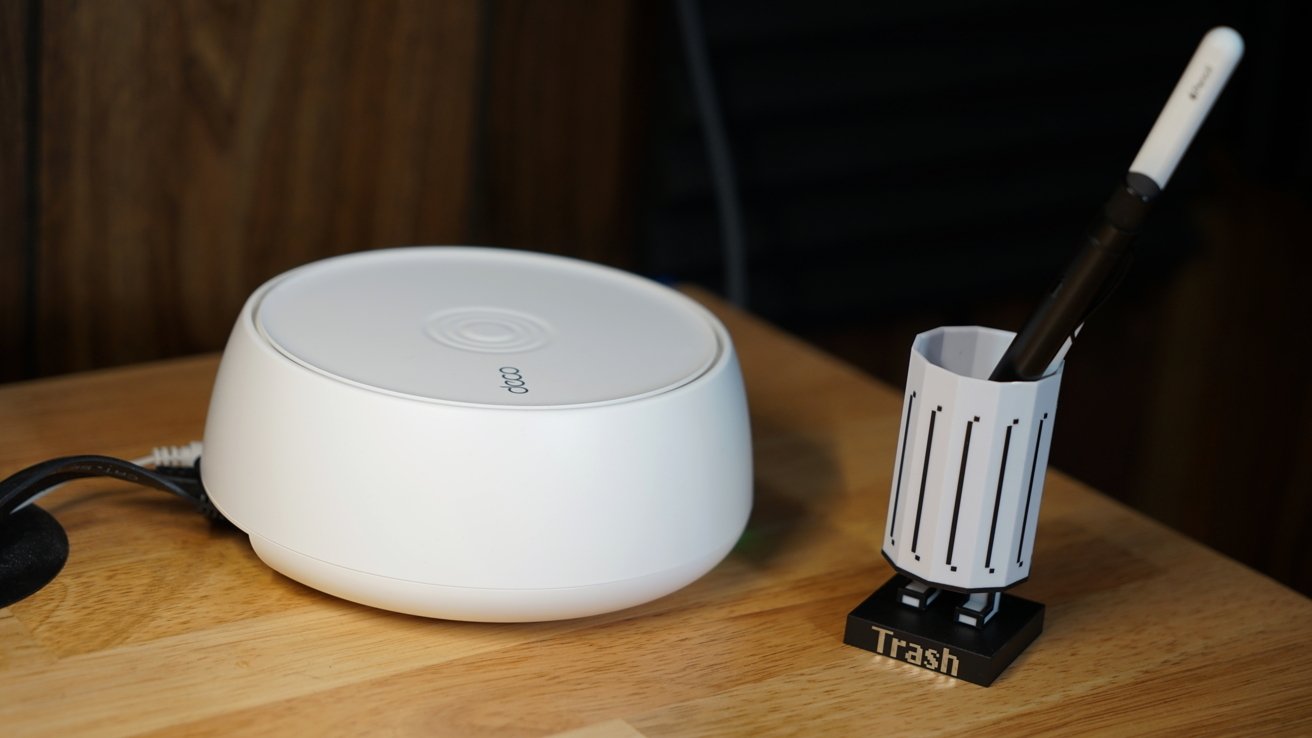 White circular device with logo and cables on a wooden surface, next to a small trash can holder containing pens.