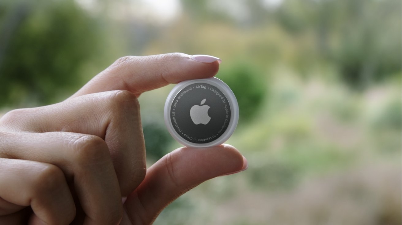 A hand holds a small round device with a logo in the center, set against a blurred natural background.