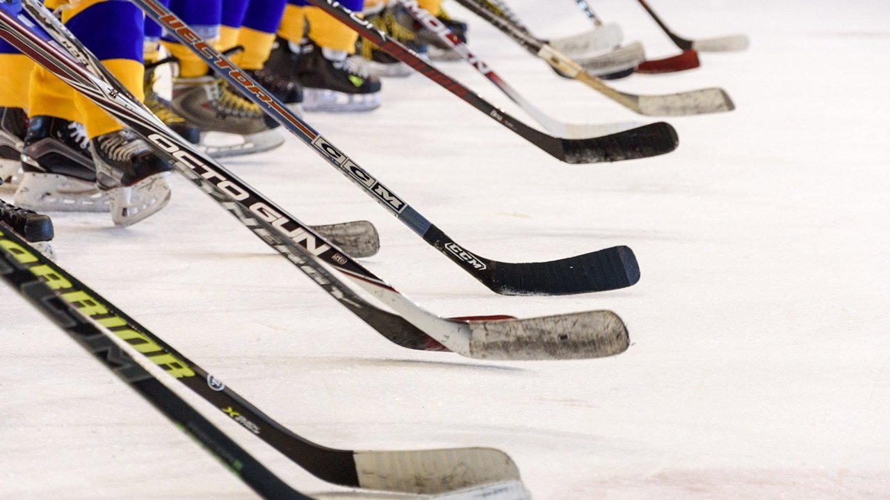 Ice hockey sticks lined up on the ice with players wearing yellow and blue uniforms and skates in the background.