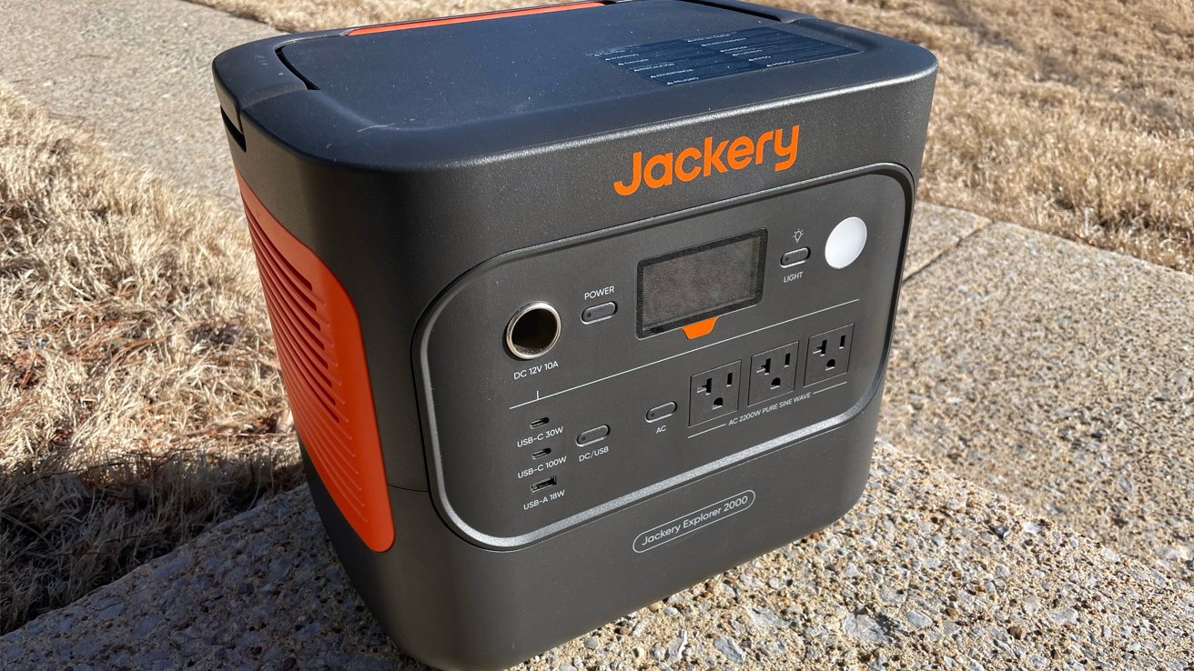 Power Station portabel dengan beberapa port dan tampilan digital, berlabel 'Jackery Explorer 2000,' bertumpu di permukaan beton di luar ruangan, dikelilingi oleh rumput kering.