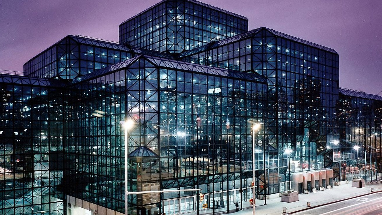 Modern glass and steel building at night with illuminated interior and exterior lights, tall geometric structures, and reflections under a purple sky.