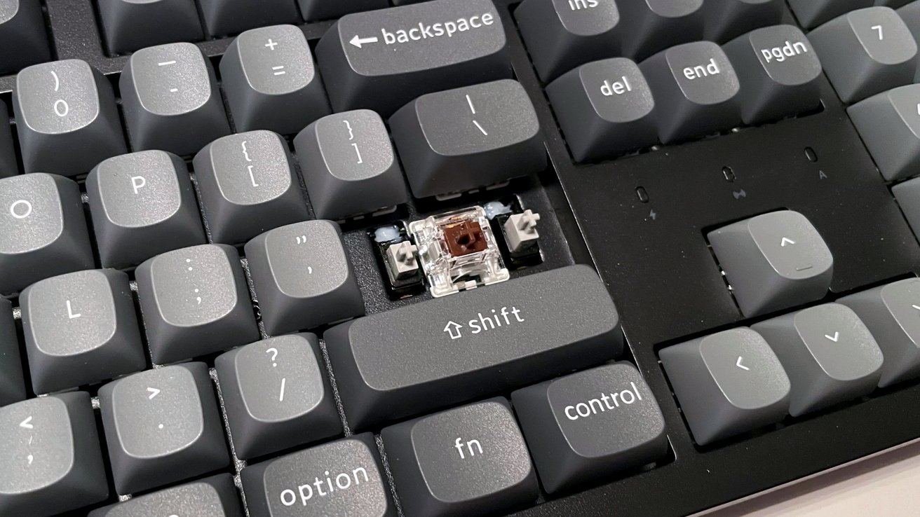 Computer keyboard with a missing keycap, exposing a brown mechanical switch beneath. Surrounding keys include backspace, shift, and arrow keys.