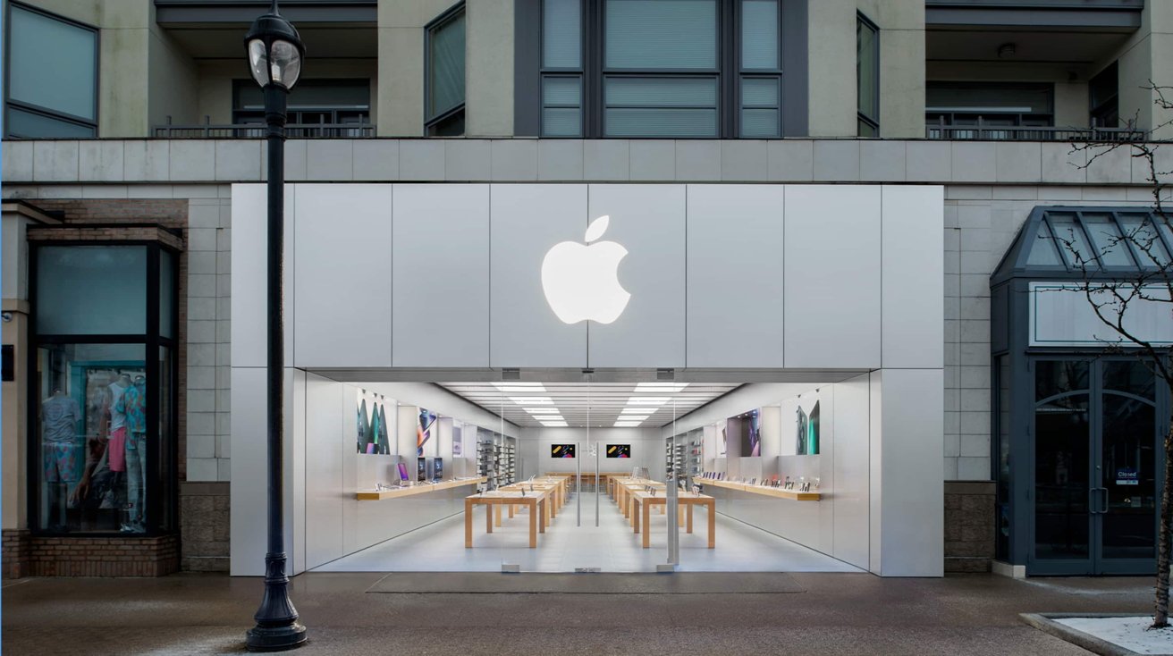 Storefront with a large lit apple logo, glass entrance, wooden tables inside, and electronic products displayed on shelves.