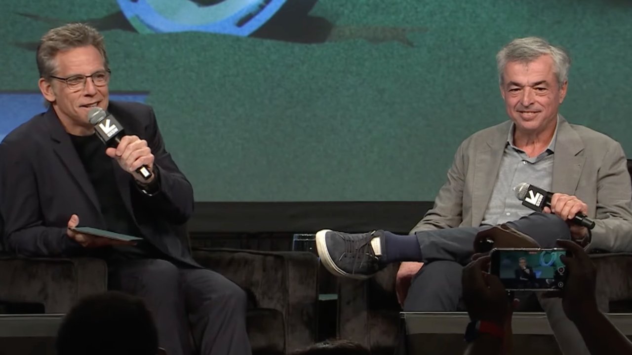 Two seated men holding microphones during a discussion, one in a black outfit and glasses, the other in a gray suit, with an audience member filming them.