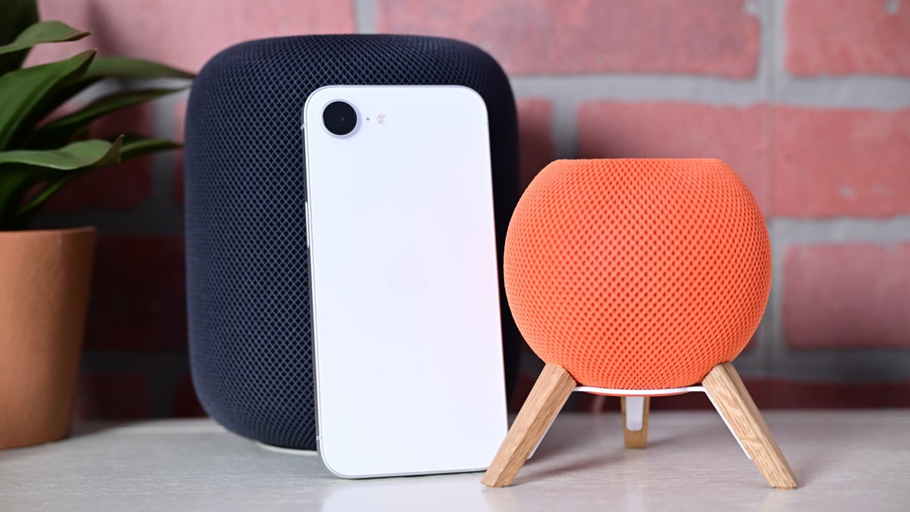 Smart speaker, white smartphone leaning against black speaker, next to an orange speaker on wooden legs, with a plant in a pot, all set against a brick wall background.