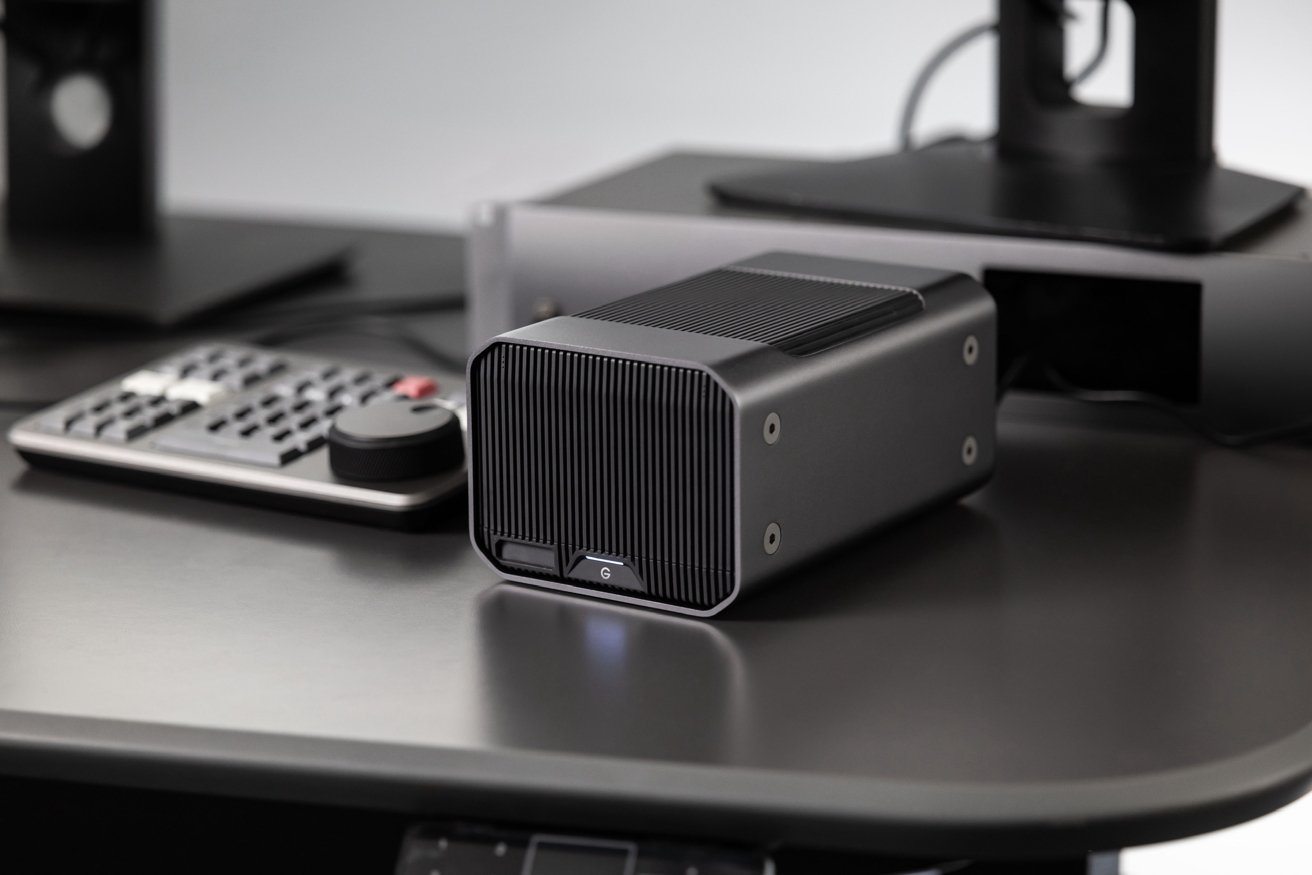 A sleek, black external hard drive is on a desk beside a computer monitor stand and a keyboard with a large knob.