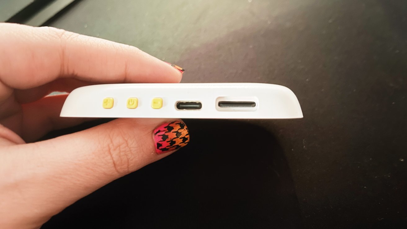 Hand holding a small white device with three yellow buttons, two ports, and patterned fingernails.