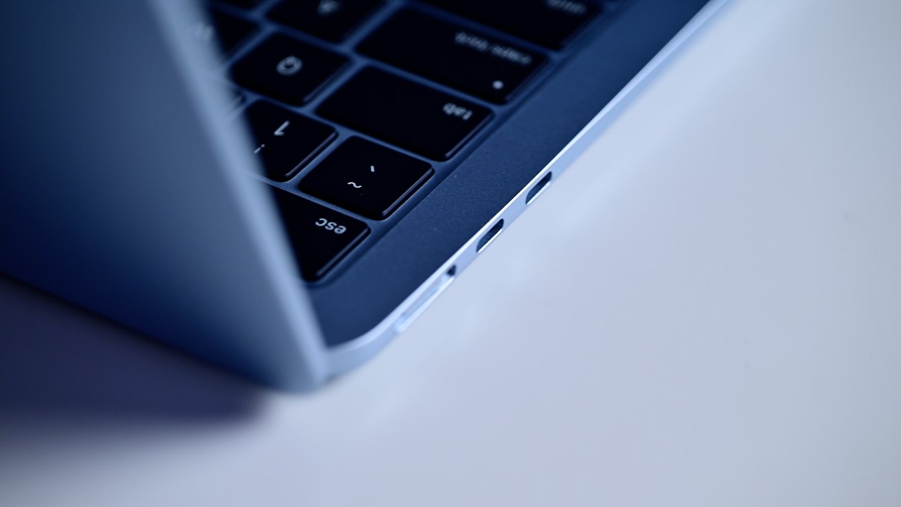 Partial view of a laptop keyboard and side ports, focusing on the escape key, with a blue-gray metallic finish on a light surface.