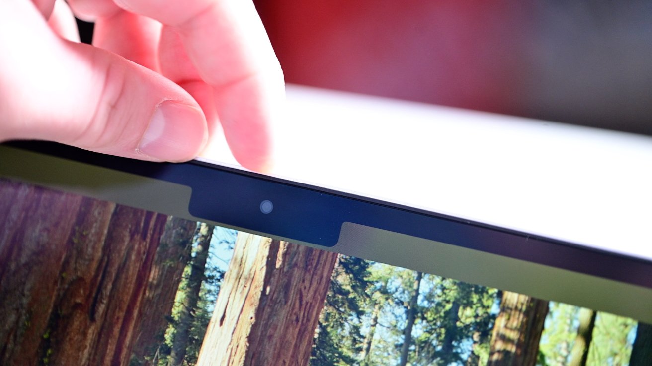 Hand adjusts the privacy cover on a webcam above a laptop screen displaying a forest scene.