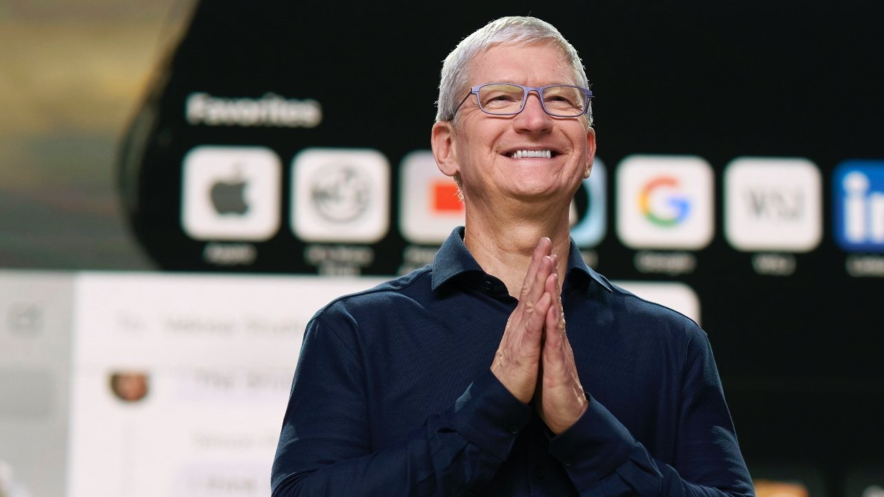 Smiling person with glasses, clasped hands, wearing a dark shirt. Blurred background features large app icons, including Apple and Google, on a screen.