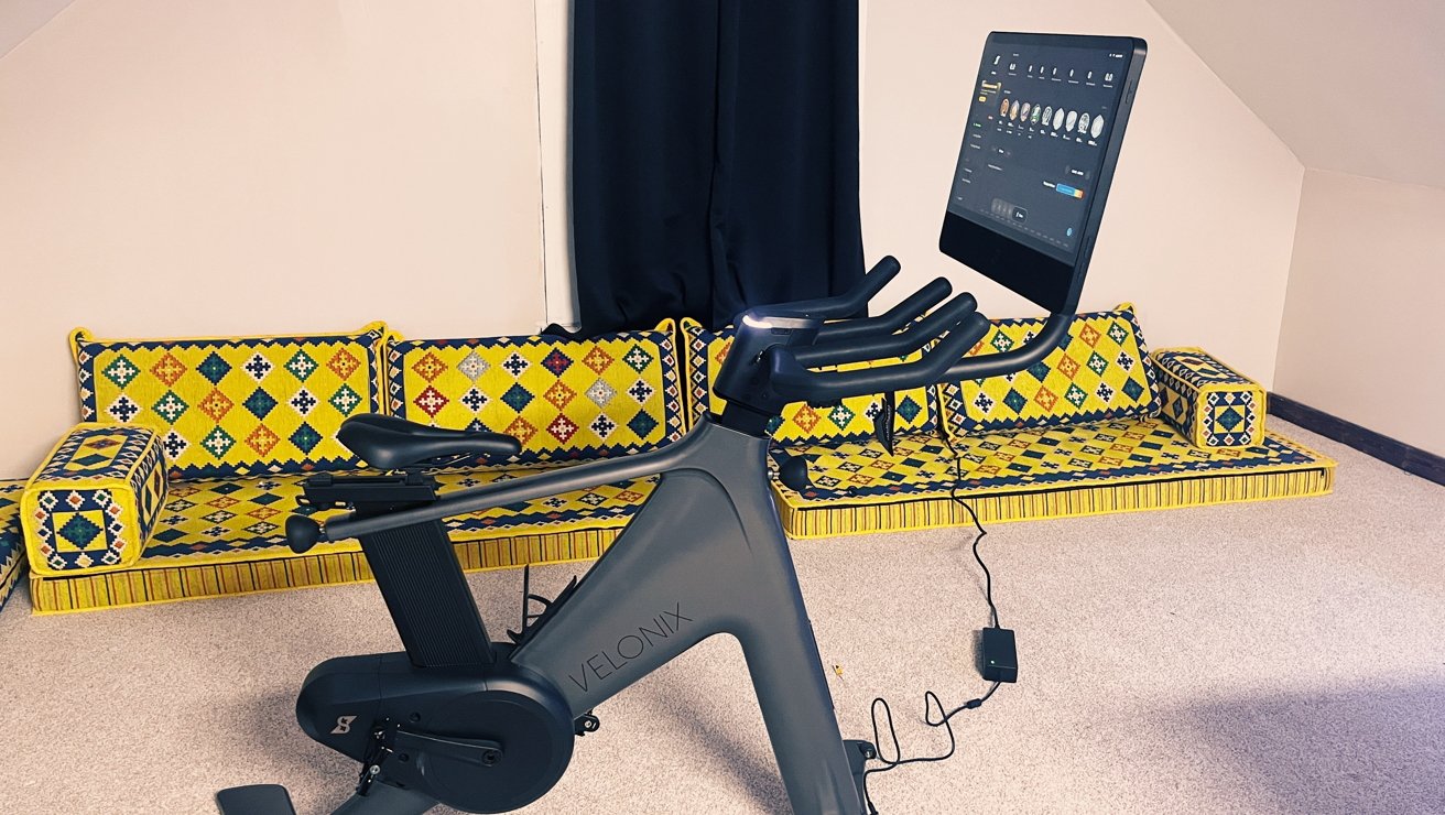 Stationary bike with digital screen in front of a colorful, patterned yellow seating area against a neutral wall.