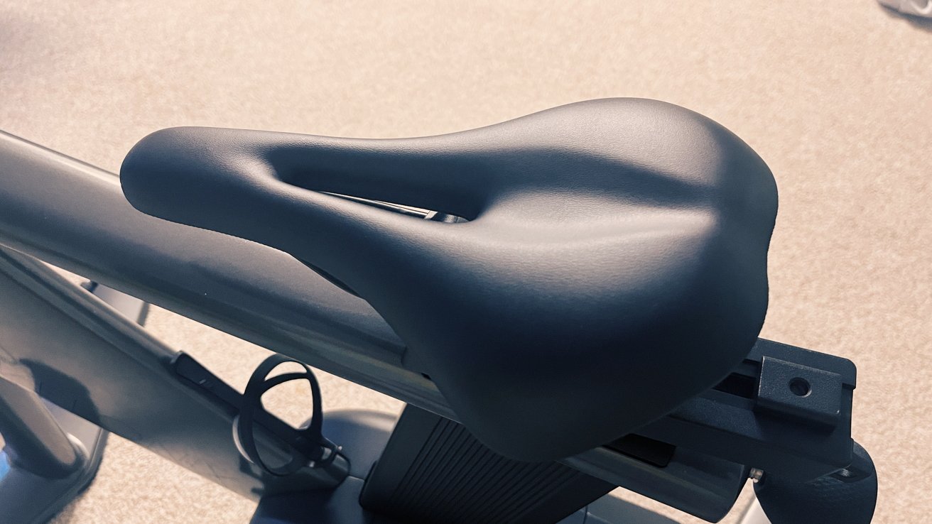 Close-up of a black bicycle saddle on a metallic bike frame, set against a carpeted floor background.