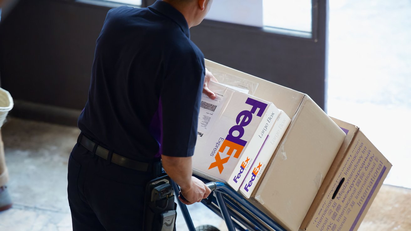 Person in uniform pushing a hand truck loaded with large FedEx boxes, inside a building entrance.