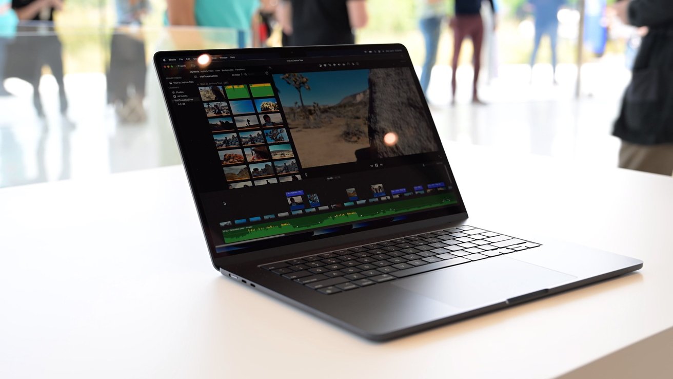 MacBook Air displaying video editing software on a desk, with several people and blurred background visible.