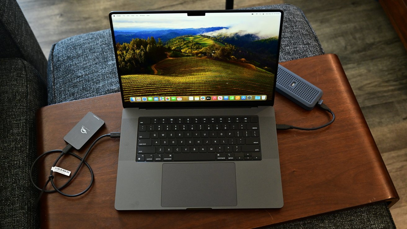 MacBook Pro di atas meja kayu menampilkan wallpaper lansekap, dengan dua drive eksternal terhubung, dikelilingi oleh furnitur kain abu -abu.