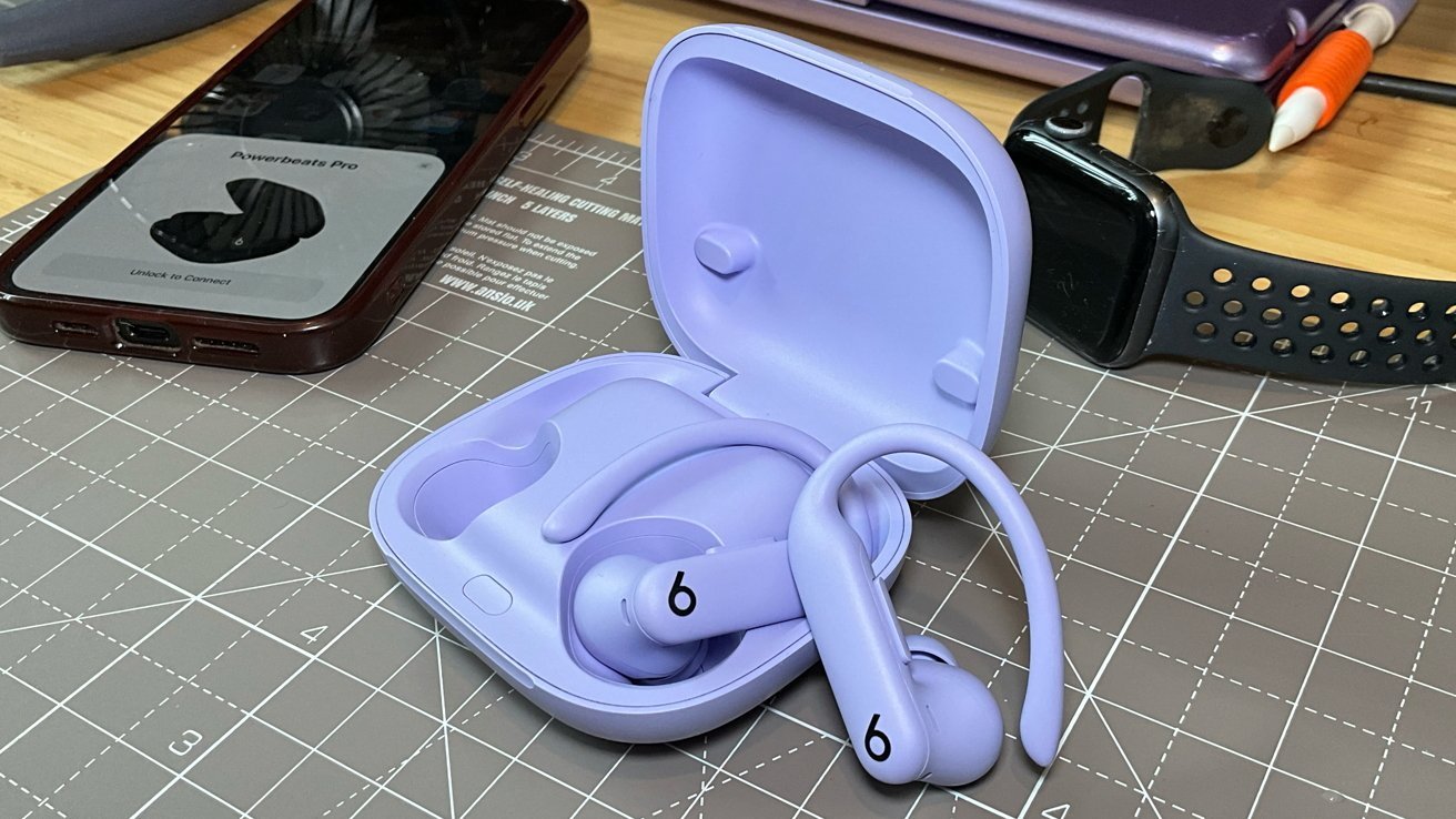 Purple wireless earbuds with ear hooks in an open charging case on a grid mat, with a smartwatch, smartphone, and pens nearby.