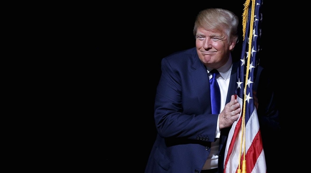 Man in a suit smiling, hugging an American flag against a dark background.