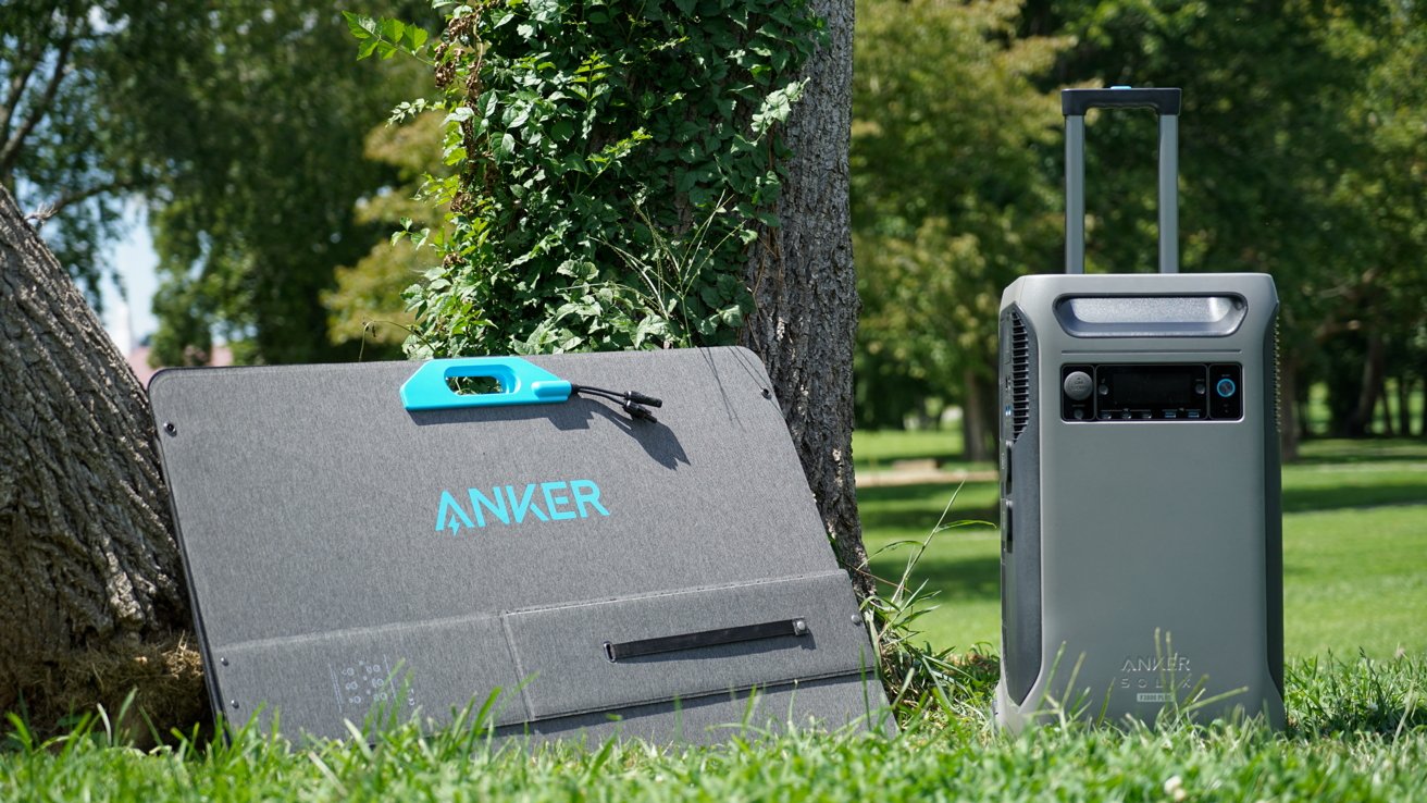 Portable solar panel and power station on grass, leaning against a tree with greenery and trees in the background.