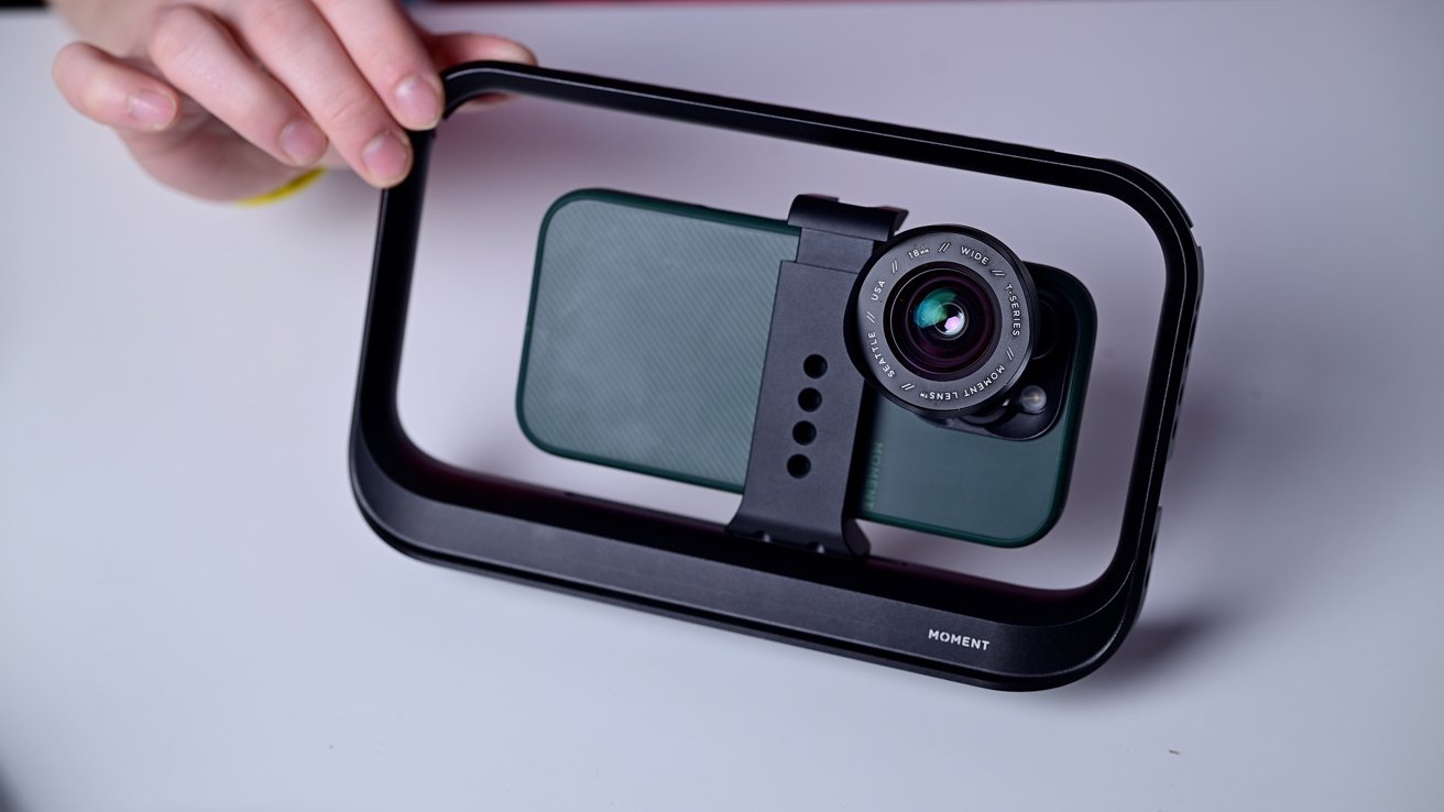 A smartphone with a camera attachment inside a black handheld frame, held by a person's hand on a white table.