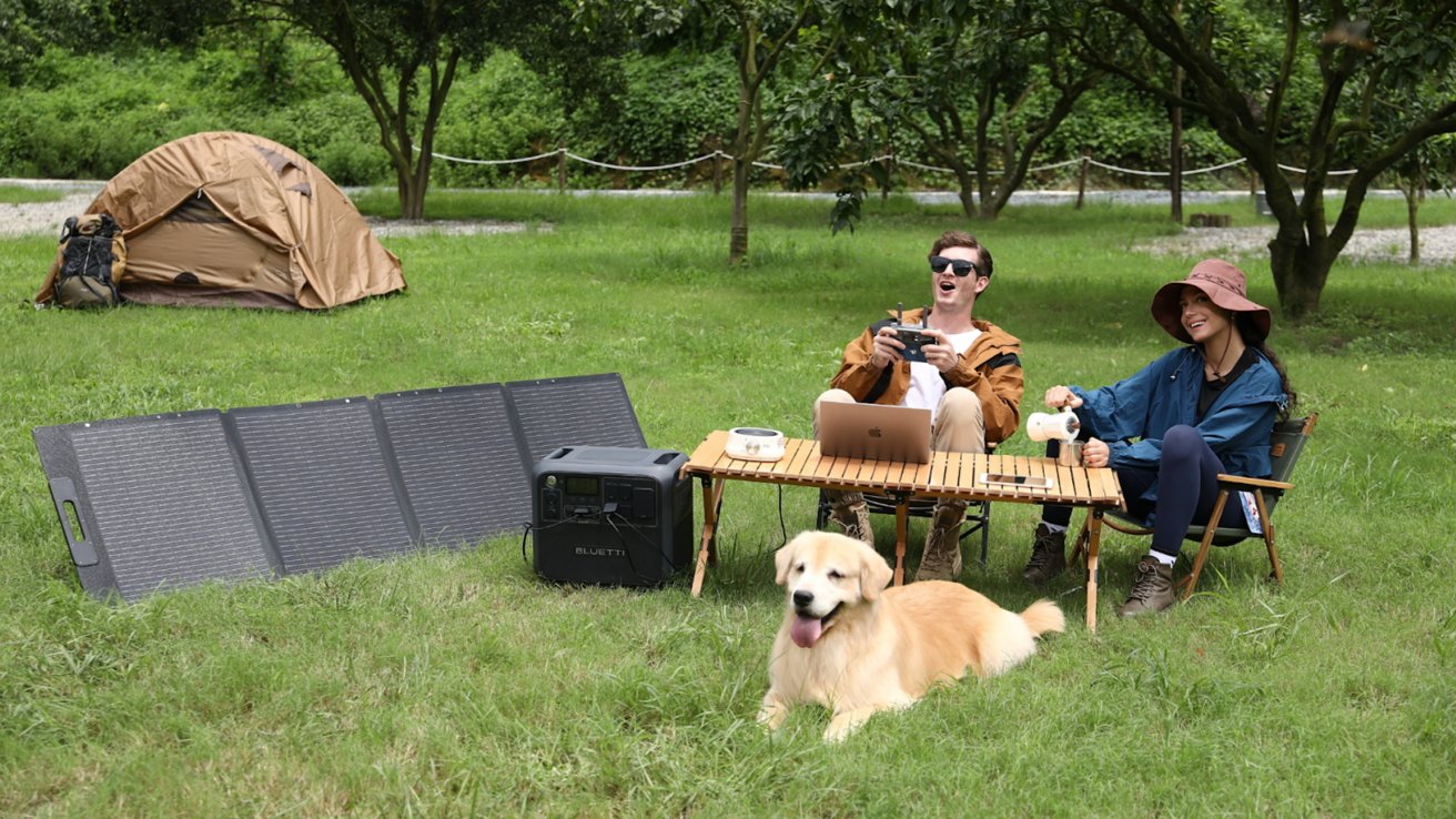 Two people enjoy camping with a dog, sitting by a table with a laptop, solar panel, and portable power station near a tent in a grassy area surrounded by trees.