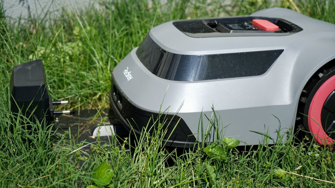 A robotic lawn mower in green grass approaches a charging station with a black plug.