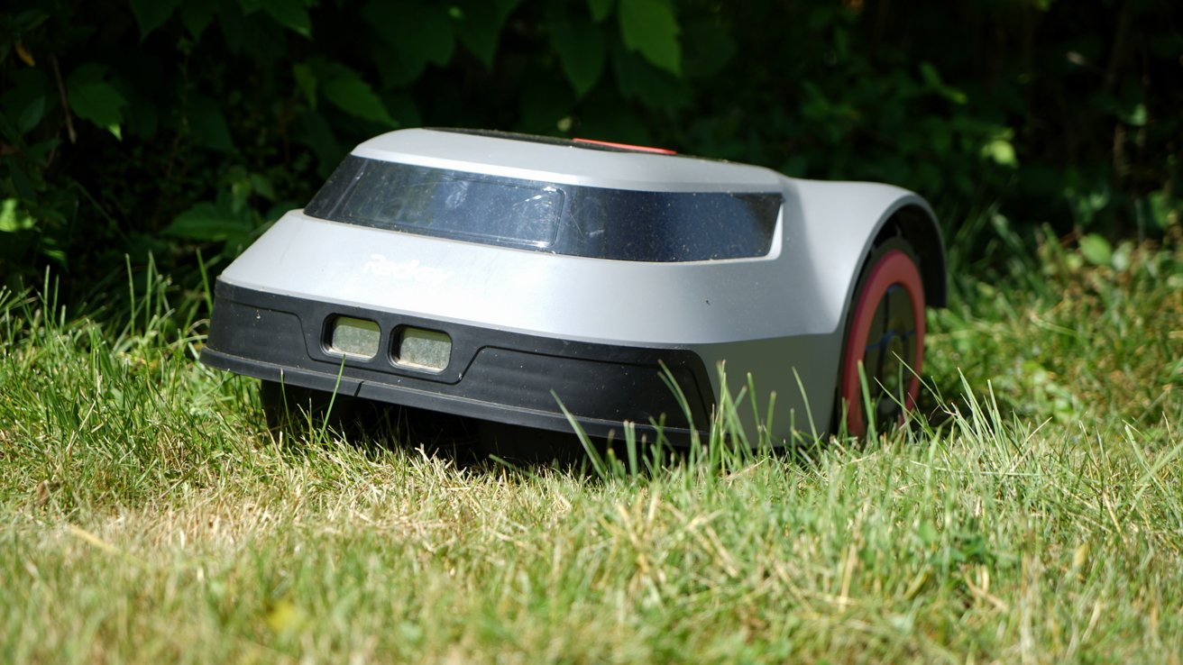 Gray robotic lawn mower on grass with a backdrop of green foliage.