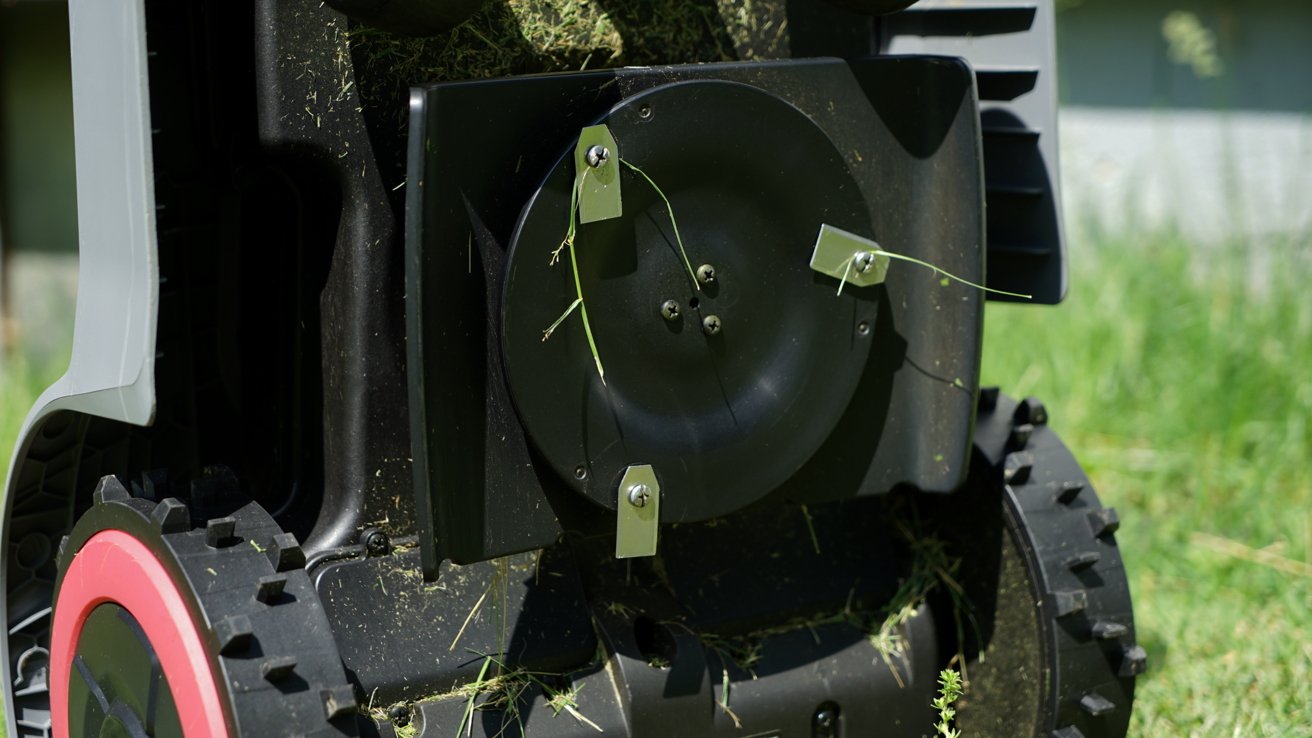 Close-up of a lawnmower's underside, showing blades and grass-covered components with red and black wheels on a sunny grassy background.