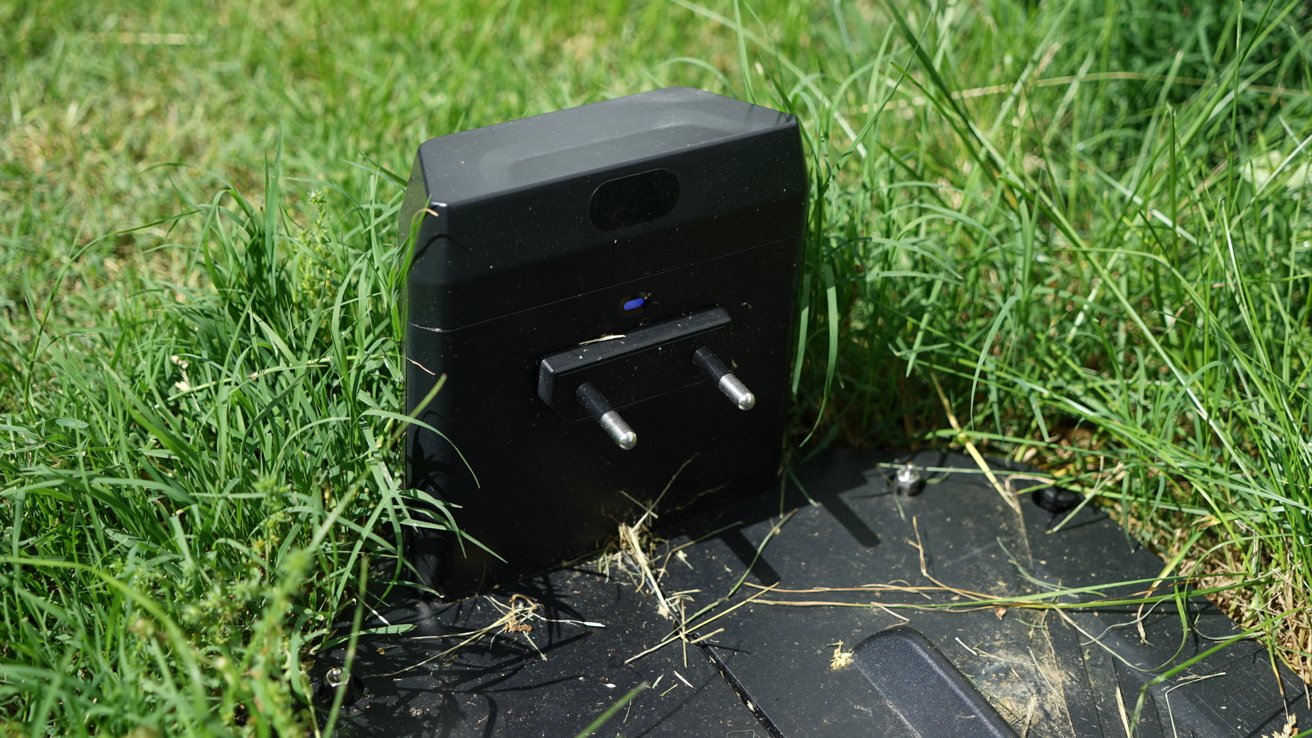 A robotic lawn mower charging station partially covered in grass, featuring a black casing and two prongs, sitting on a base.