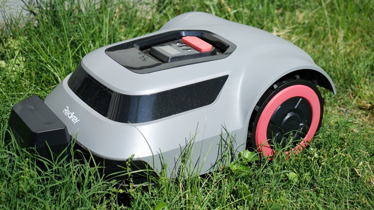 A silver robotic lawnmower with pink wheels sits on green grass, featuring a black panel on top with a red button.