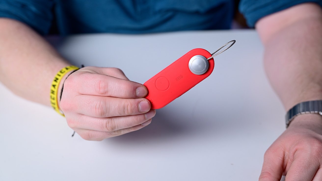 Hand holding a red keychain tracker with a round silver button and a wristband visible on the wrist.