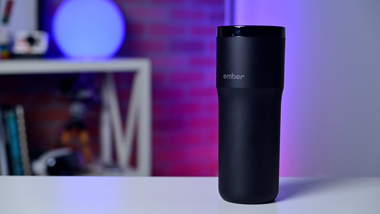 A black travel mug labeled 'ember' on a white table, with a blurred purple and red-lit background featuring shelves and books.