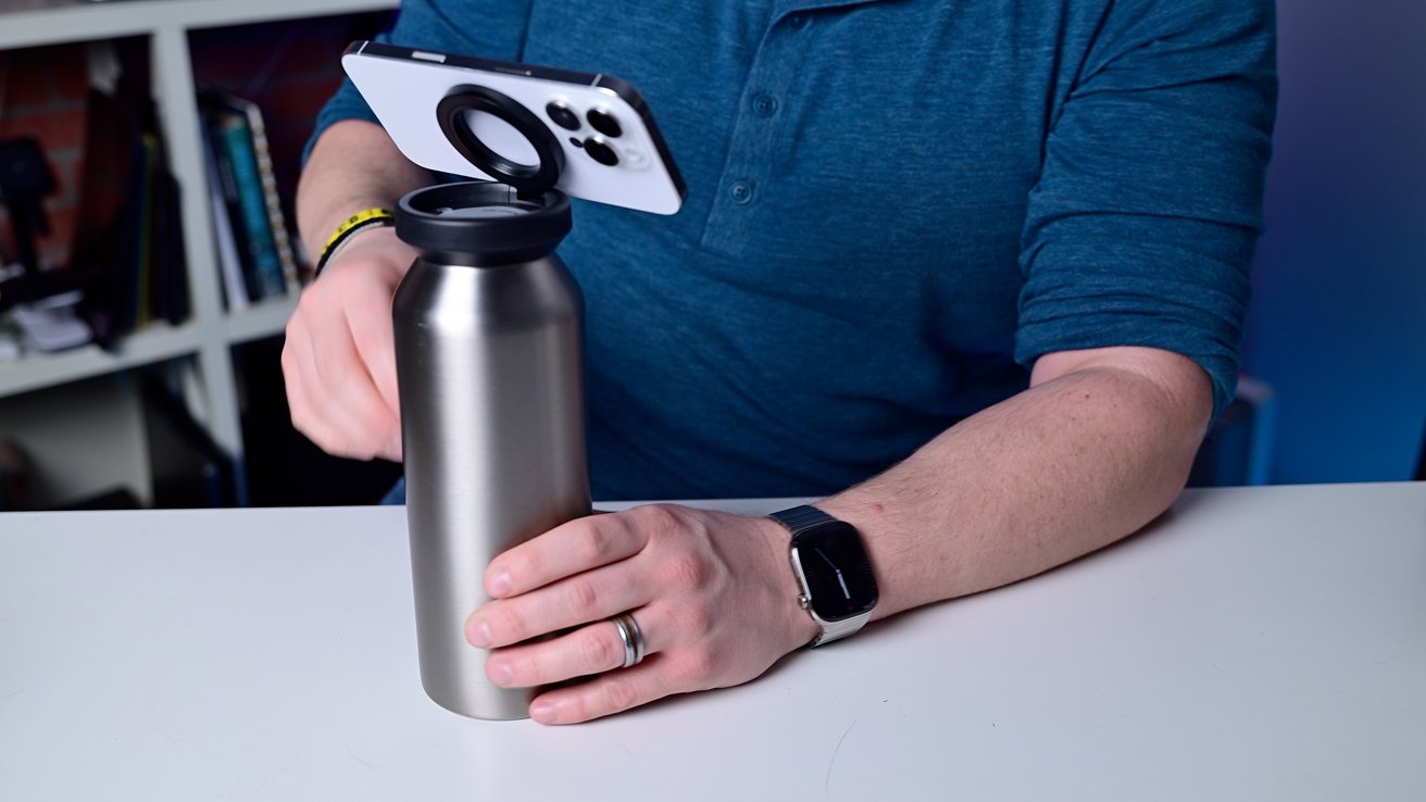 Person wearing a smartwatch holds a metal bottle on a table. A phone is attached to the bottle cap with a ring holder. Books are visible in the background.