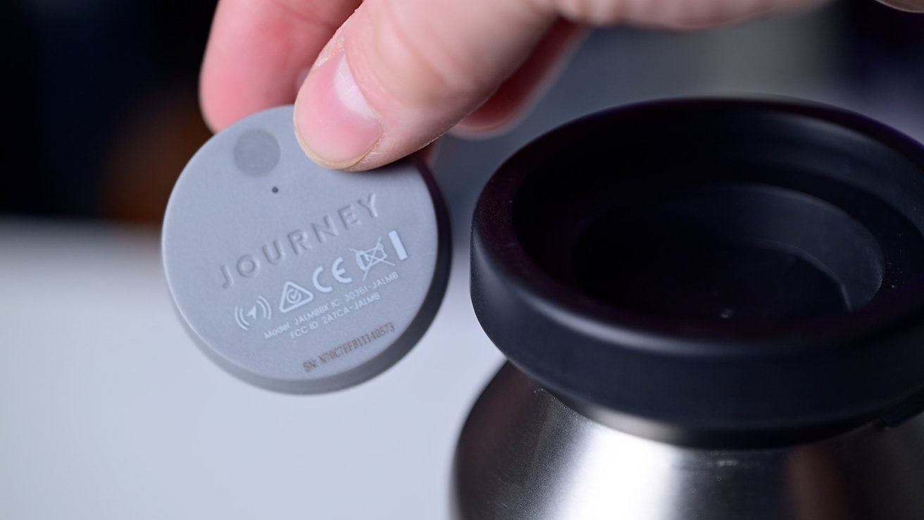 A hand holding a gray circular electronic tag labeled Journey near a metal container.